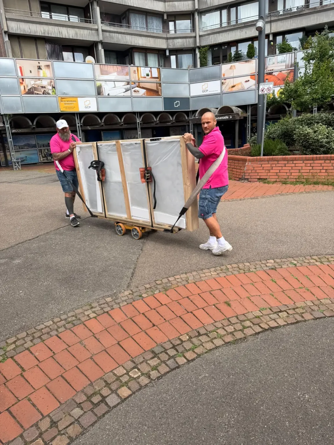 Zwei Mitarbeiter in pinken Shirts bewegen ein verkistetes Klavier auf einem Rollbrett